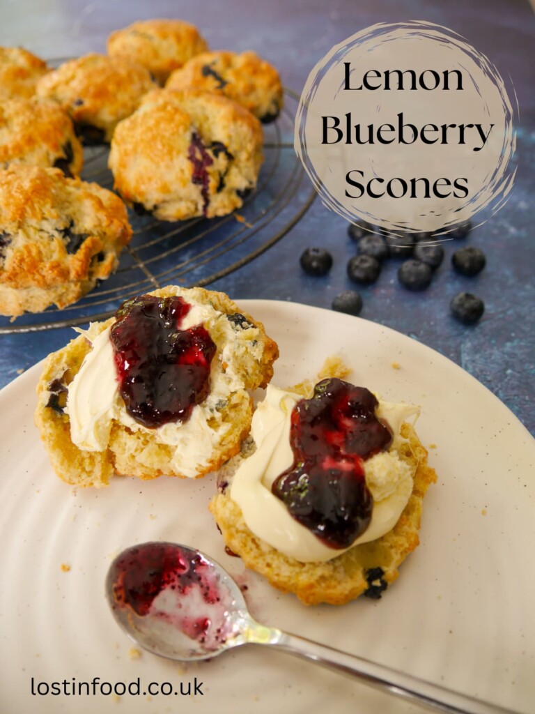 Pinnable image with recipe title and a plate of lemon and blueberry scones, topped with clotted cream and blueberry jam. A wire rack of scones is set alongside.