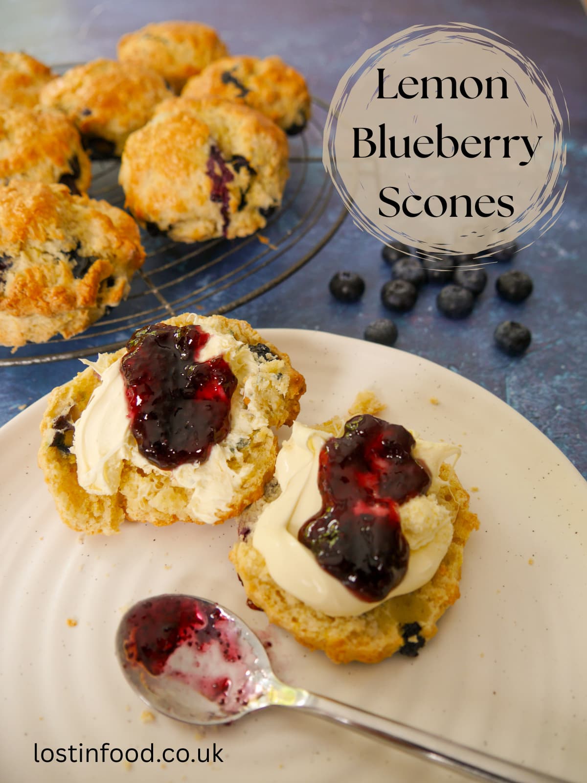 Pinnable image with recipe title and a plate of lemon and blueberry scones, topped with clotted cream and blueberry jam. A wire rack of scones is set alongside.