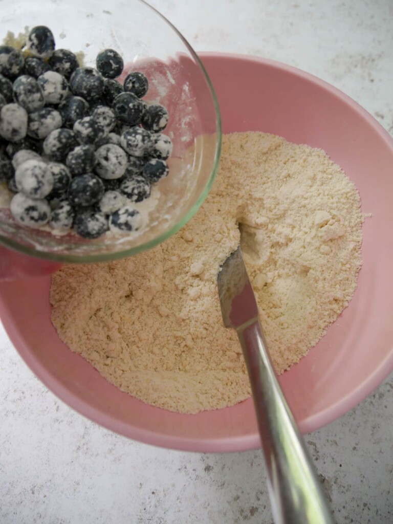 A bowl of dry scone ingredients with blueberries being added. The blueberries have a light coating of flour on them.
