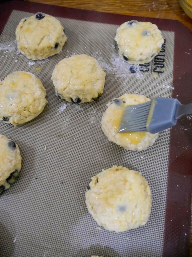 A lined baking sheet with fresh cut lemon and blueberry scones being brushed with egg wash.
