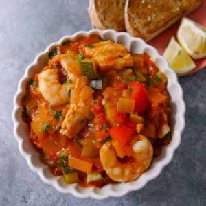 A bowl of Mediterranean fish soup served with slices of toasted sourdough and wedges of lemon.