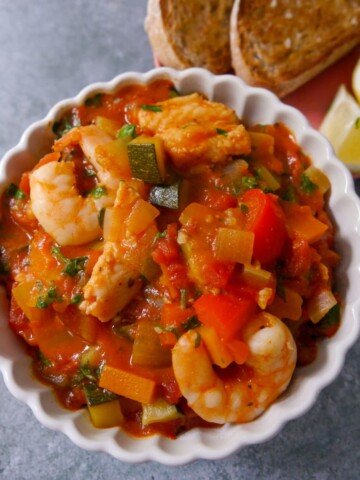 A bowl of Mediterranean fish soup served with slices of toasted sourdough and wedges of lemon.