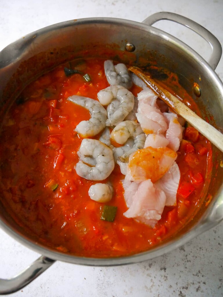 A large soup pan of cooked Mediterranean fish soup with chunks of fresh white fish and raw prawns being added.