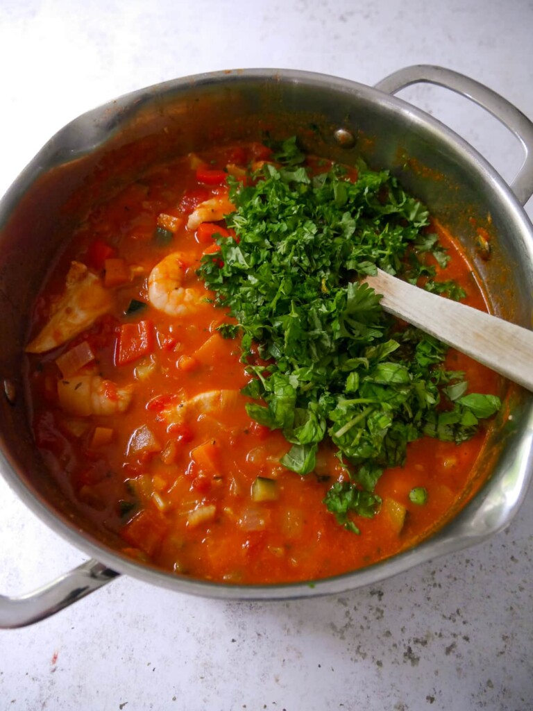 A large soup pan of cooked Mediterranean fish soup with freshly chopped basil and parsley being added.