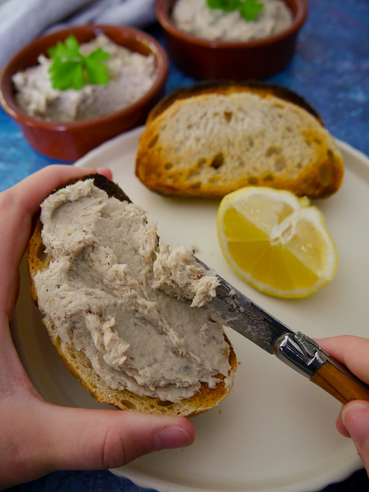 A slice of toast being spread with smoked mackerel pate, with bowls of pate and wedges of lemon set alongside.