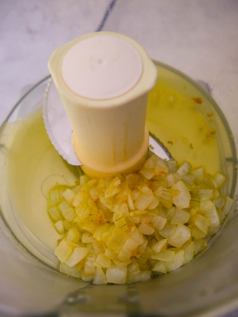 Bowl of a food processor filled with melted butter and cooked chopped onions and garlic.