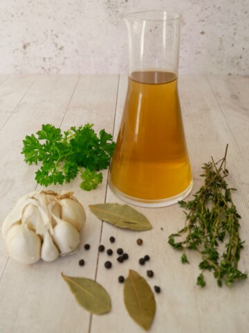 A jug of homemade vegetable stock, with fresh herbs and aromatics set alongside.