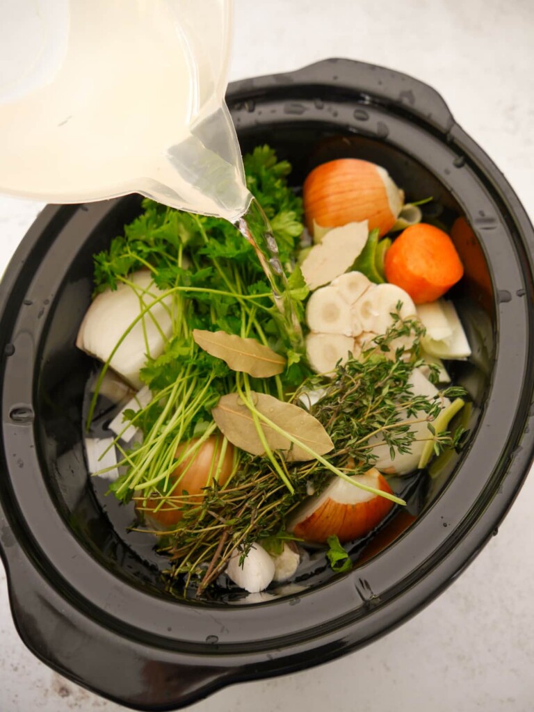 A slow cooker bowl filled with fresh chopped vegetables and herbs with water being poured over.