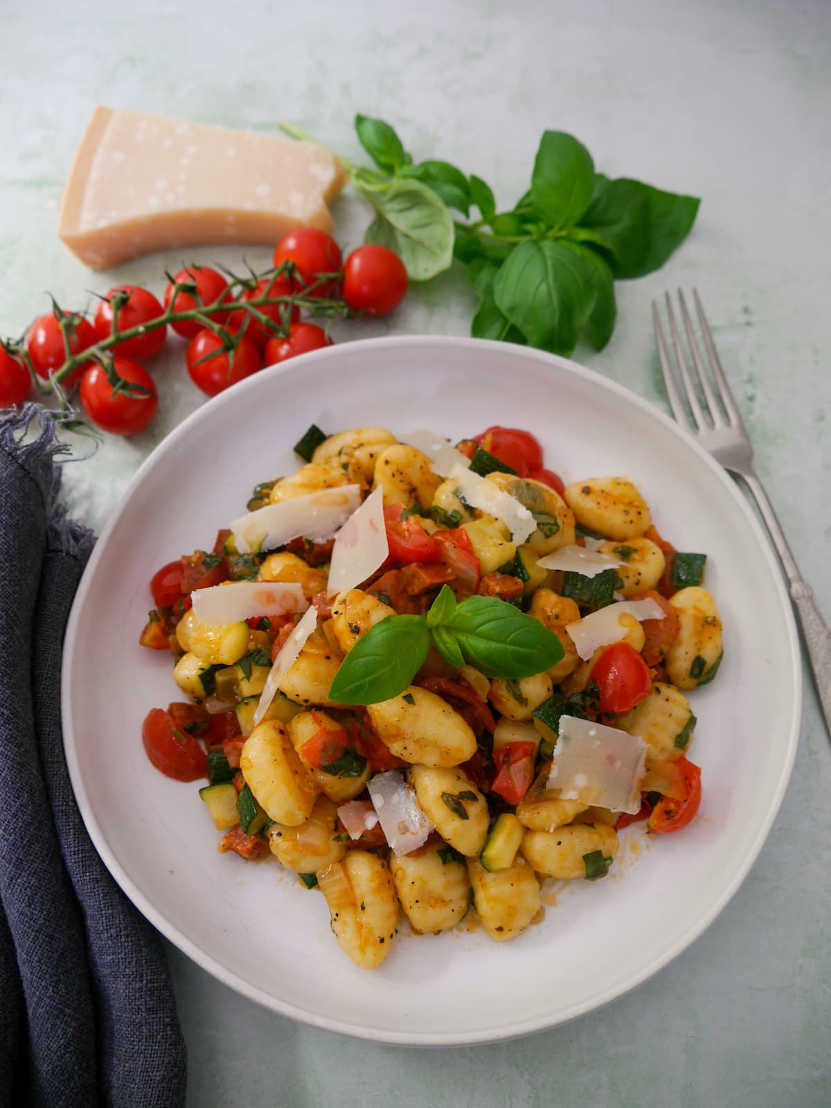 A white bowl filled with chorizo gnocchi with sauteed vegetables, garnished with parmesan cheese and fresh basil, with basil, tomatoes and parmesan cheese set alongside.