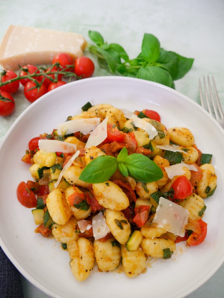 A white bowl filled with chorizo gnocchi with sauteed vegetables, garnished with parmesan cheese and fresh basil, with basil, tomatoes and parmesan cheese set alongside.