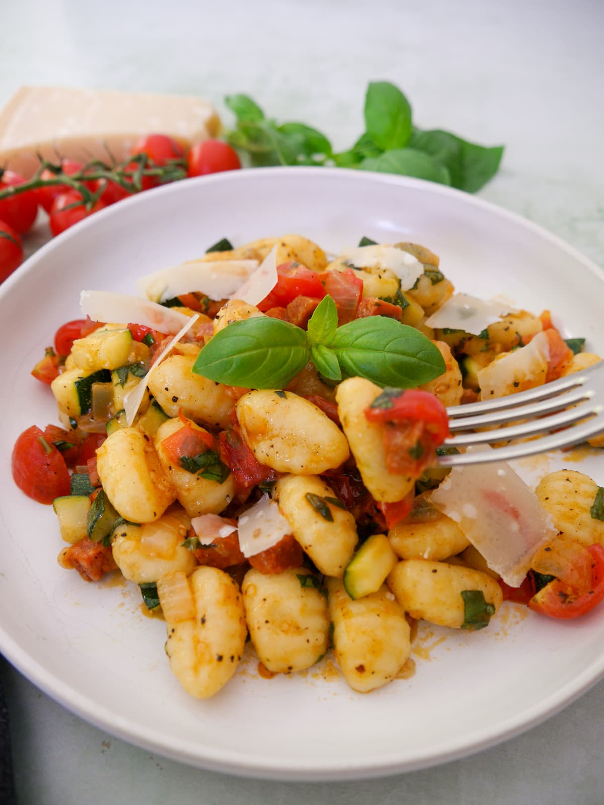 A white bowl filled with chorizo gnocchi with sauteed vegetables, garnished with parmesan cheese and fresh basil, with basil, tomatoes and parmesan cheese set alongside.
