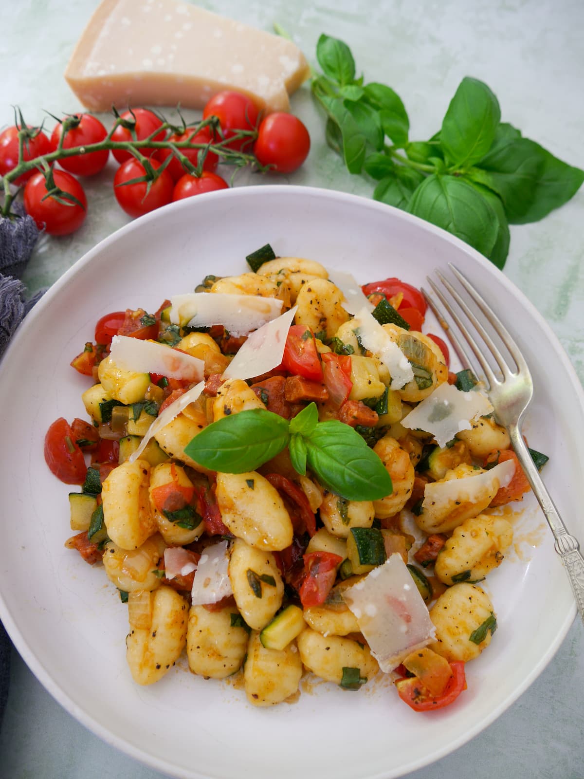 A white bowl filled with chorizo gnocchi with sauteed vegetables, garnished with parmesan cheese and fresh basil, with basil, tomatoes and parmesan cheese set alongside.