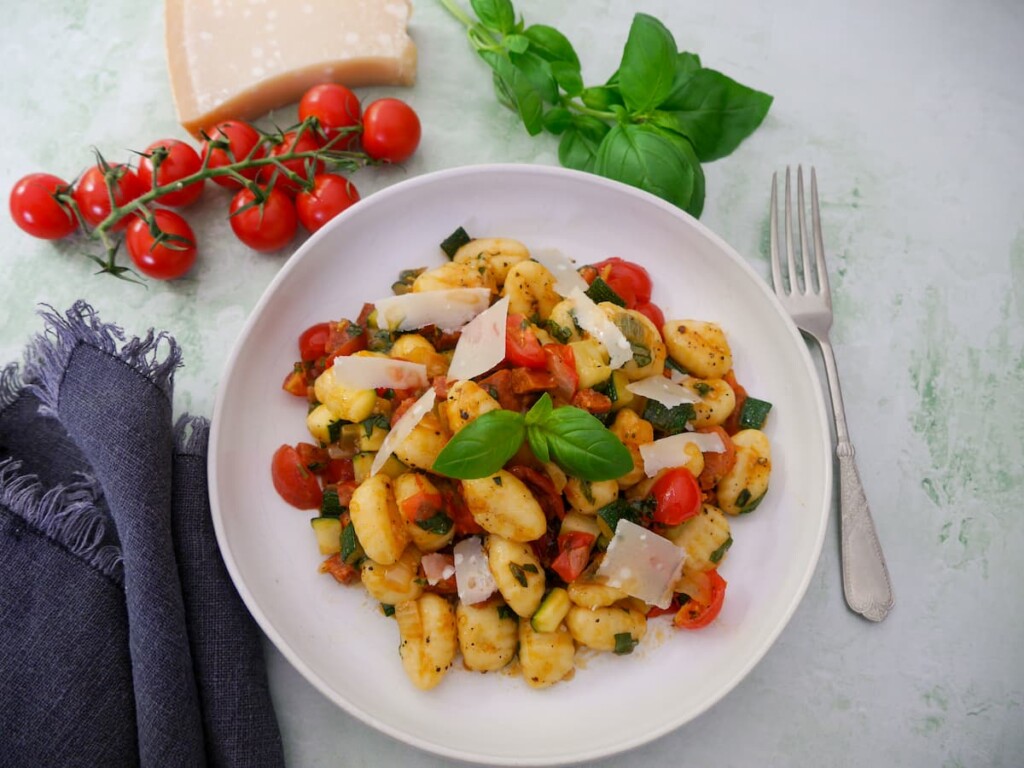 A white bowl filled with chorizo gnocchi with sauteed vegetables, garnished with parmesan cheese and fresh basil, with basil, tomatoes and parmesan cheese set alongside.