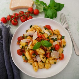 A white bowl filled with chorizo gnocchi with sauteed vegetables, garnished with parmesan cheese and fresh basil, with basil, tomatoes and parmesan cheese set alongside.
