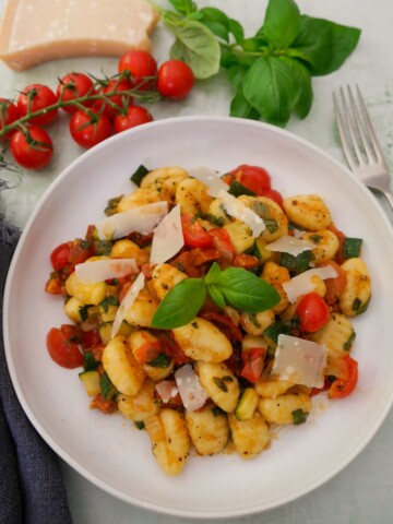 A white bowl filled with chorizo gnocchi with sauteed vegetables, garnished with parmesan cheese and fresh basil, with basil, tomatoes and parmesan cheese set alongside.
