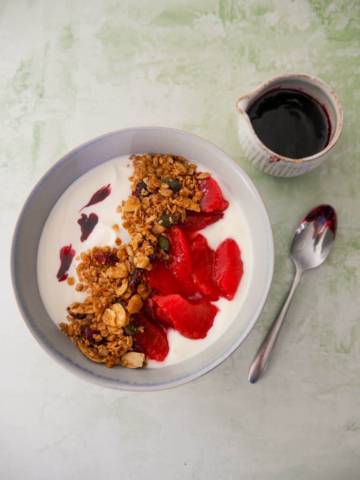 A bowl of Greek yogurt topped with mulled plums and granola, with a jug of mulled plum syrup set alongside.