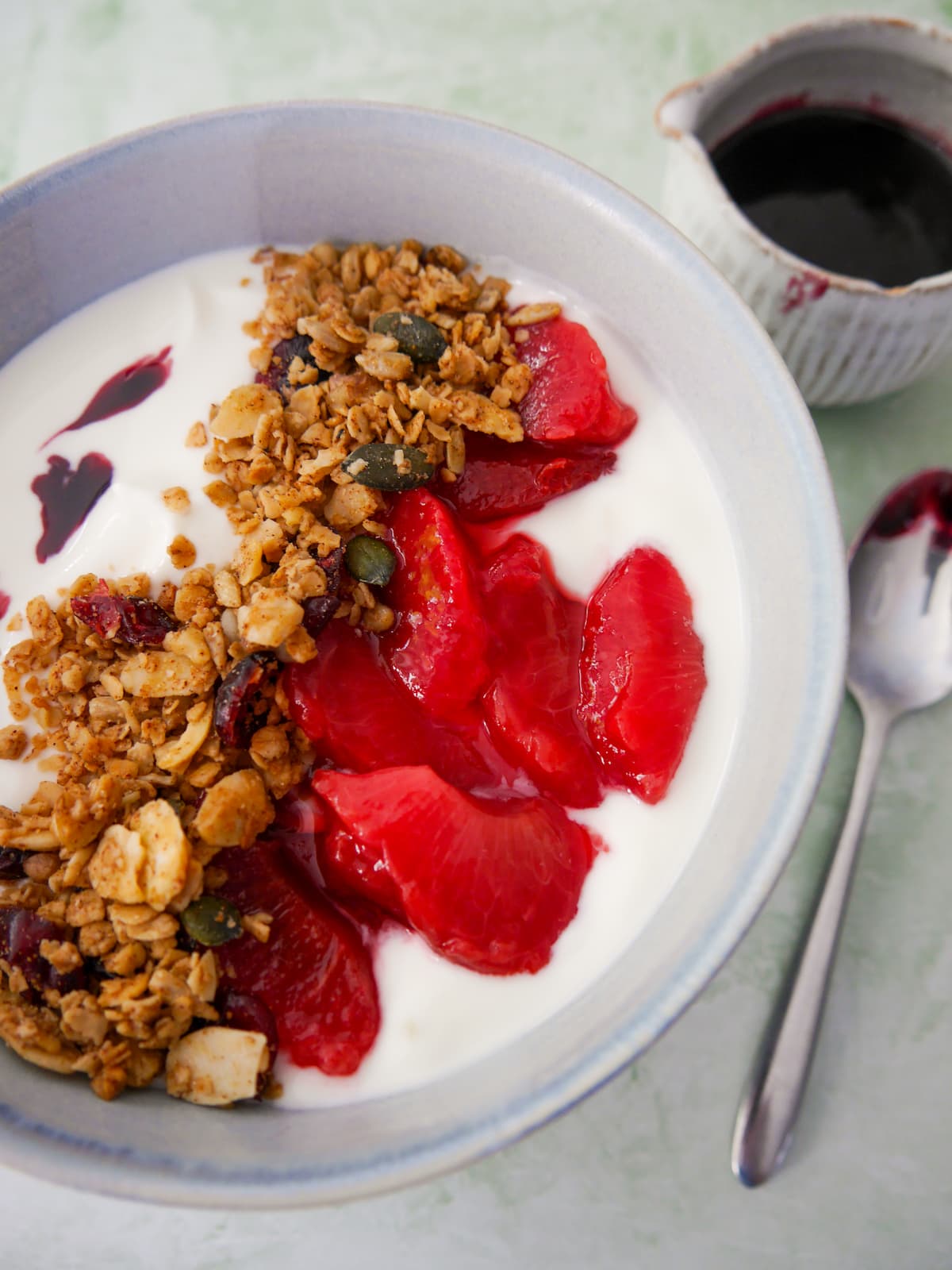 A bowl of Greek yogurt topped with mulled plums and granola, with a jug of mulled plum syrup set alongside.