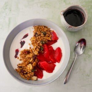 A bowl of Greek yogurt topped with mulled plums and granola, with a jug of mulled plum syrup set alongside.