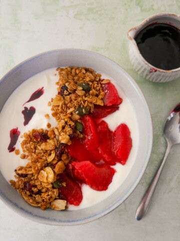 A bowl of Greek yogurt topped with mulled plums and granola, with a jug of mulled plum syrup set alongside.