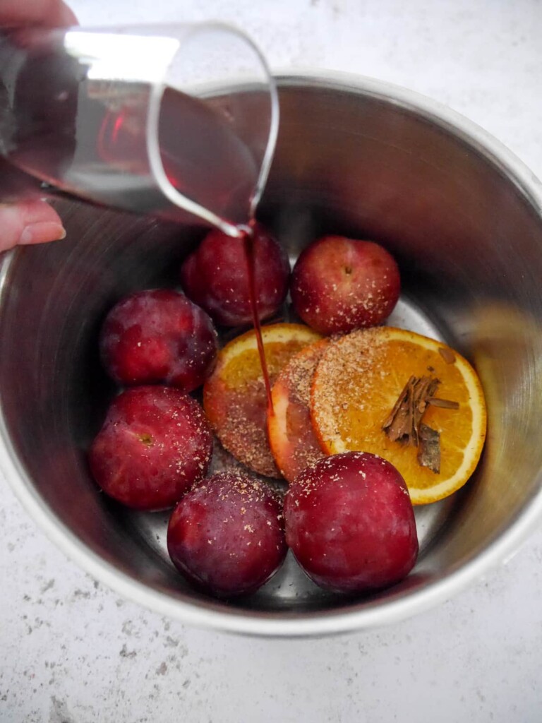 Red wine being poured into a saucepan of plums, with sliced orange, cinnamon stick and grated nutmeg.