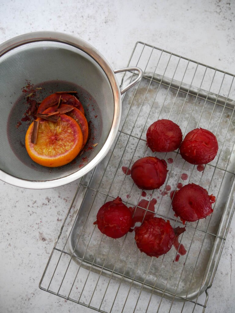 A sieve of mulled plum liquor set over a bowl, with the mulled plums set alongside on a wire rack set over a baking tray.