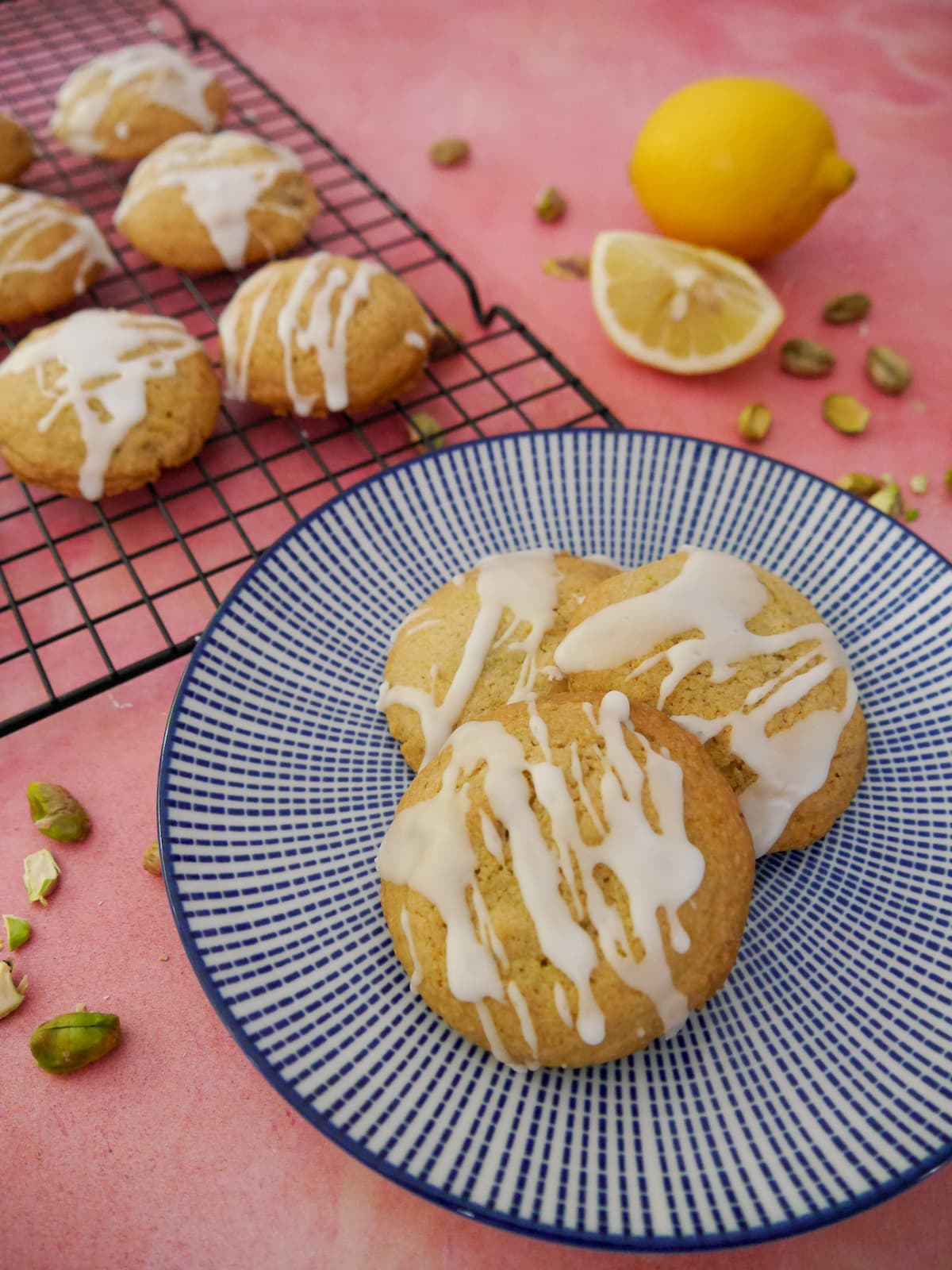 A blue and white plate topped with pistachio and lemon cookies, with more cookies set on a wire cooling rack set alongside.