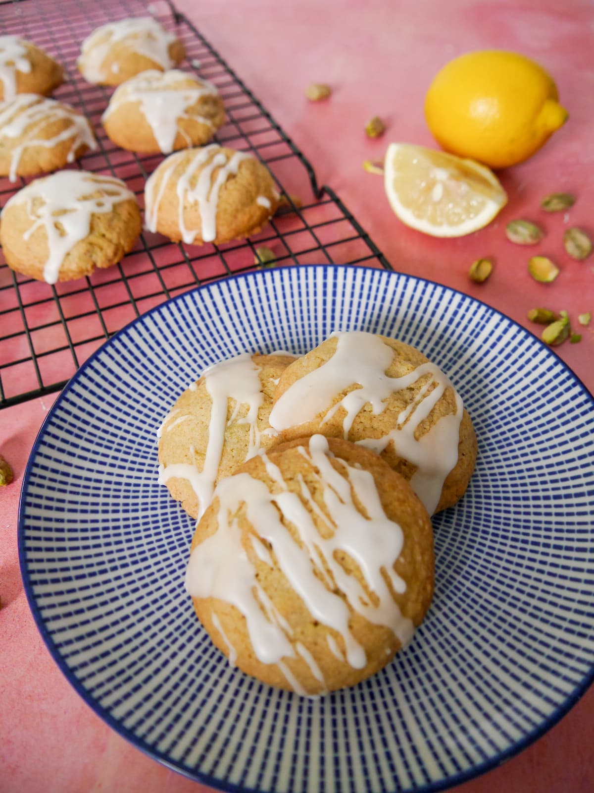 A blue and white plate topped with pistachio and lemon cookies, with more cookies set on a wire cooling rack set alongside.