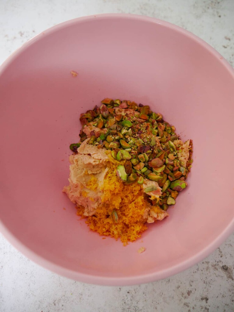 A pink mixing bowl filled with a cookie dough base with added chopped pistachio nuts and finely grated lemon zest.