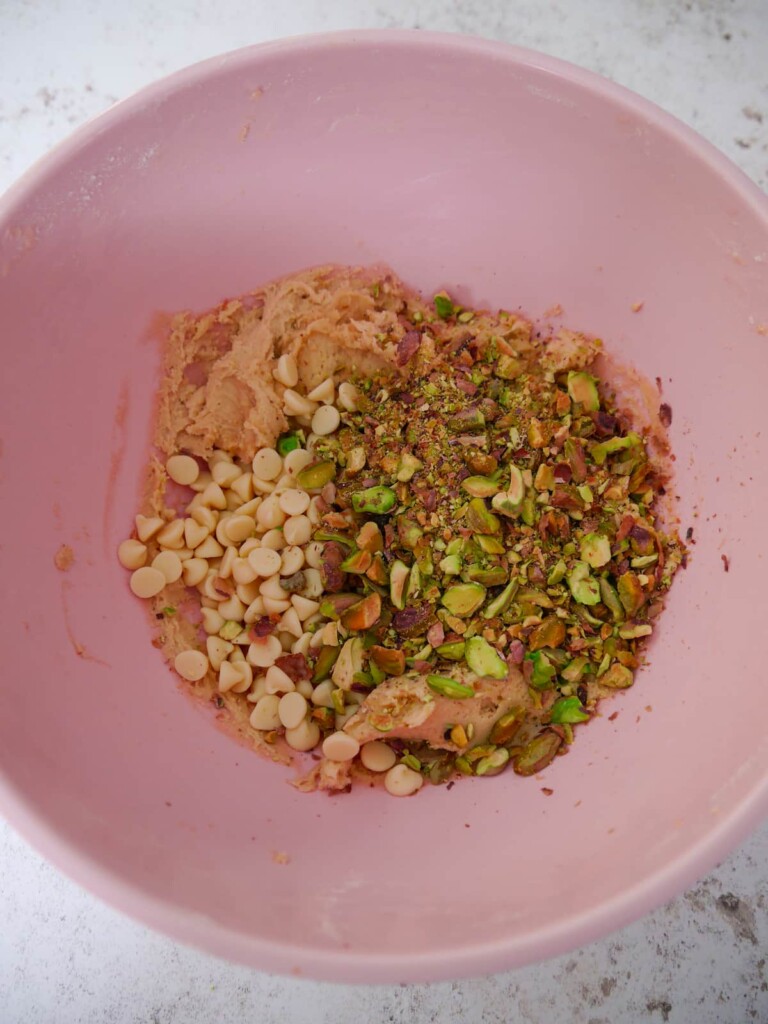A pink bowl of cookie dough with added chopped pistachio nuts and white chocolate chips.
