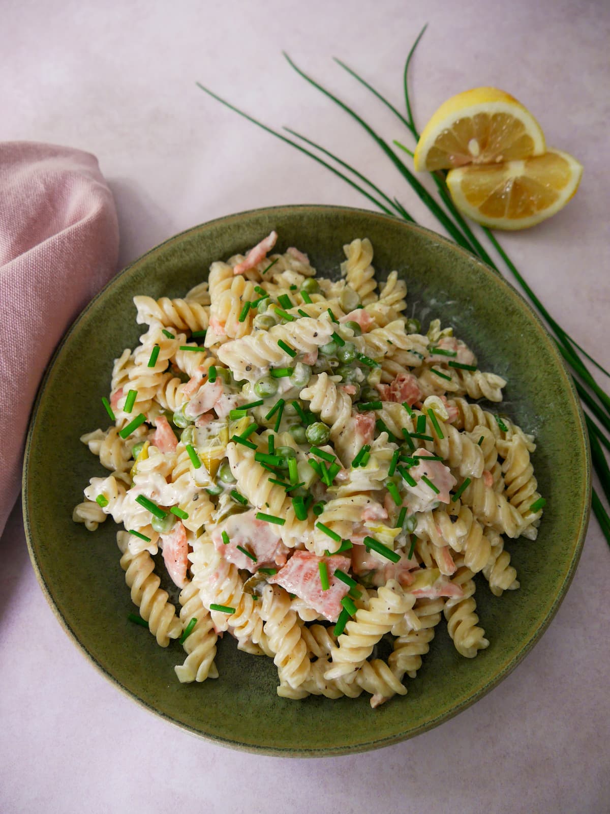 A green bowl filled with smoked salmon pasta garnished with chopped chives with a wedge of lemon and chives set alongside.