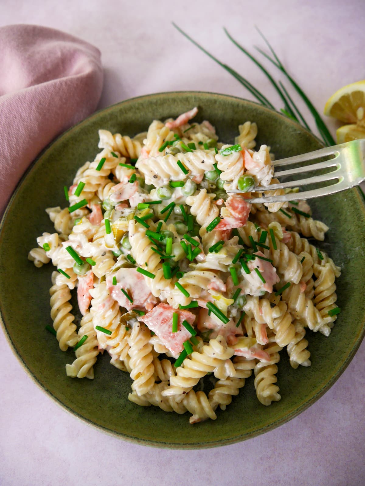 A green bowl filled with smoked salmon pasta garnished with chopped chives with a wedge of lemon and chives set alongside.