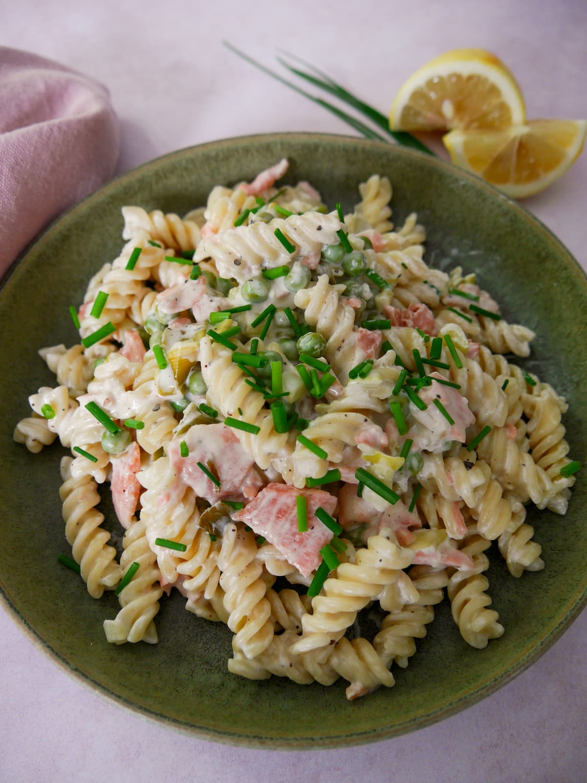 A green bowl filled with smoked salmon pasta garnished with chopped chives with a wedge of lemon and chives set alongside.