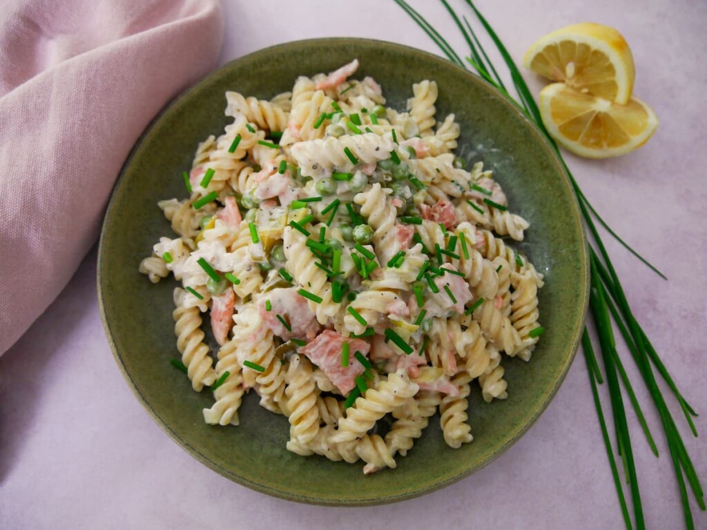 A green bowl filled with smoked salmon pasta garnished with chopped chives with a wedge of lemon and chives set alongside.