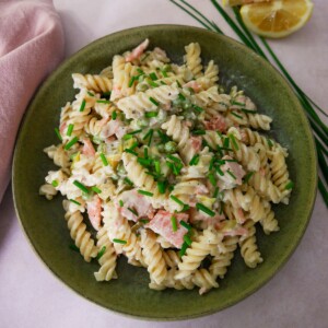 A green bowl filled with smoked salmon pasta garnished with chopped chives with a wedge of lemon and chives set alongside.