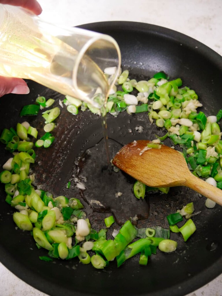 A large frying pan of sauteed spring onion and garlic with white wine being added.