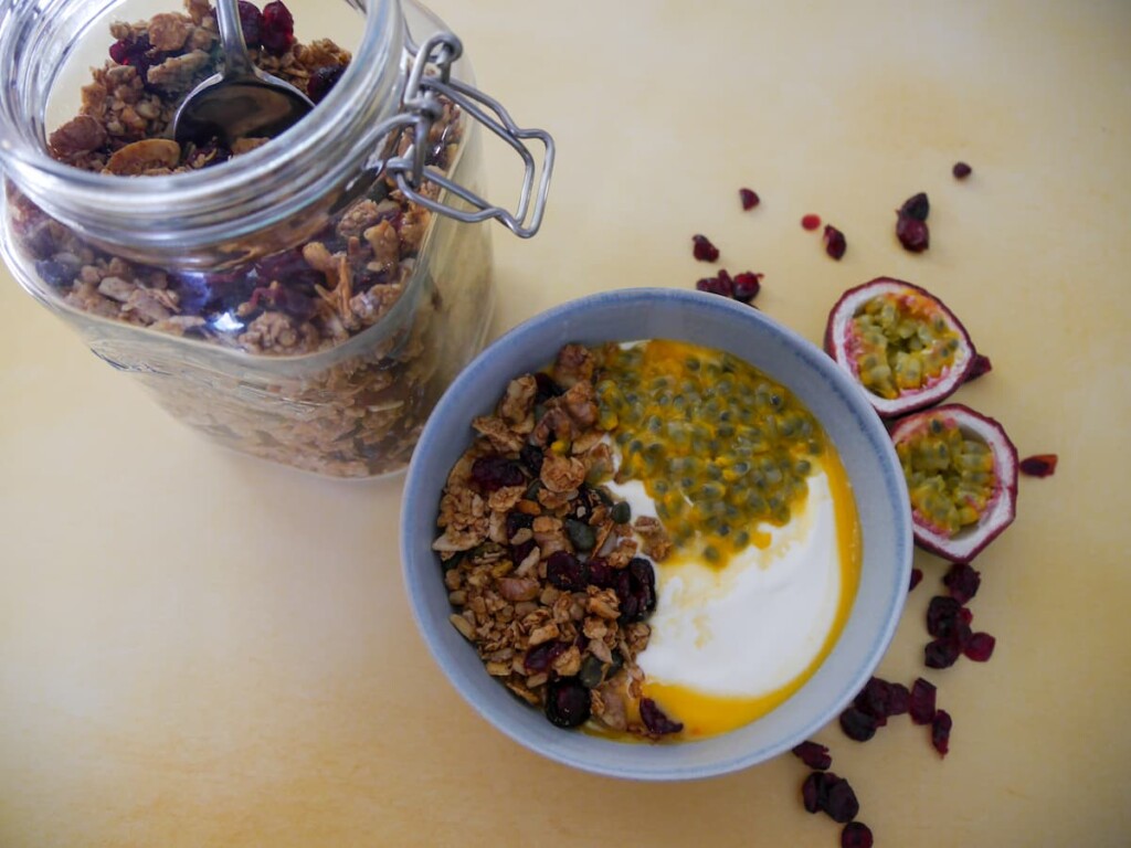 A bowl of Greek yogurt topped with spiced cranberry granola and passionfruit with a jar of granola set alongside.