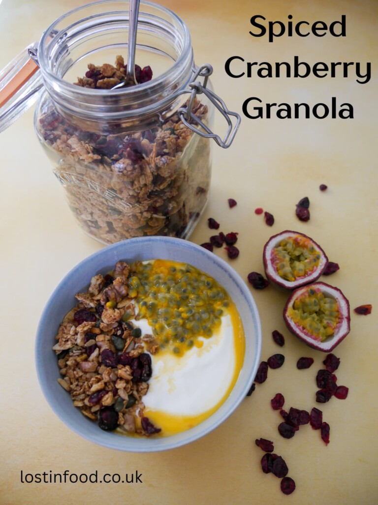 Pinnable image with recipe title and a bowl of Greek yogurt topped with spiced cranberry granola and passionfruit with a jar of granola set alongside.