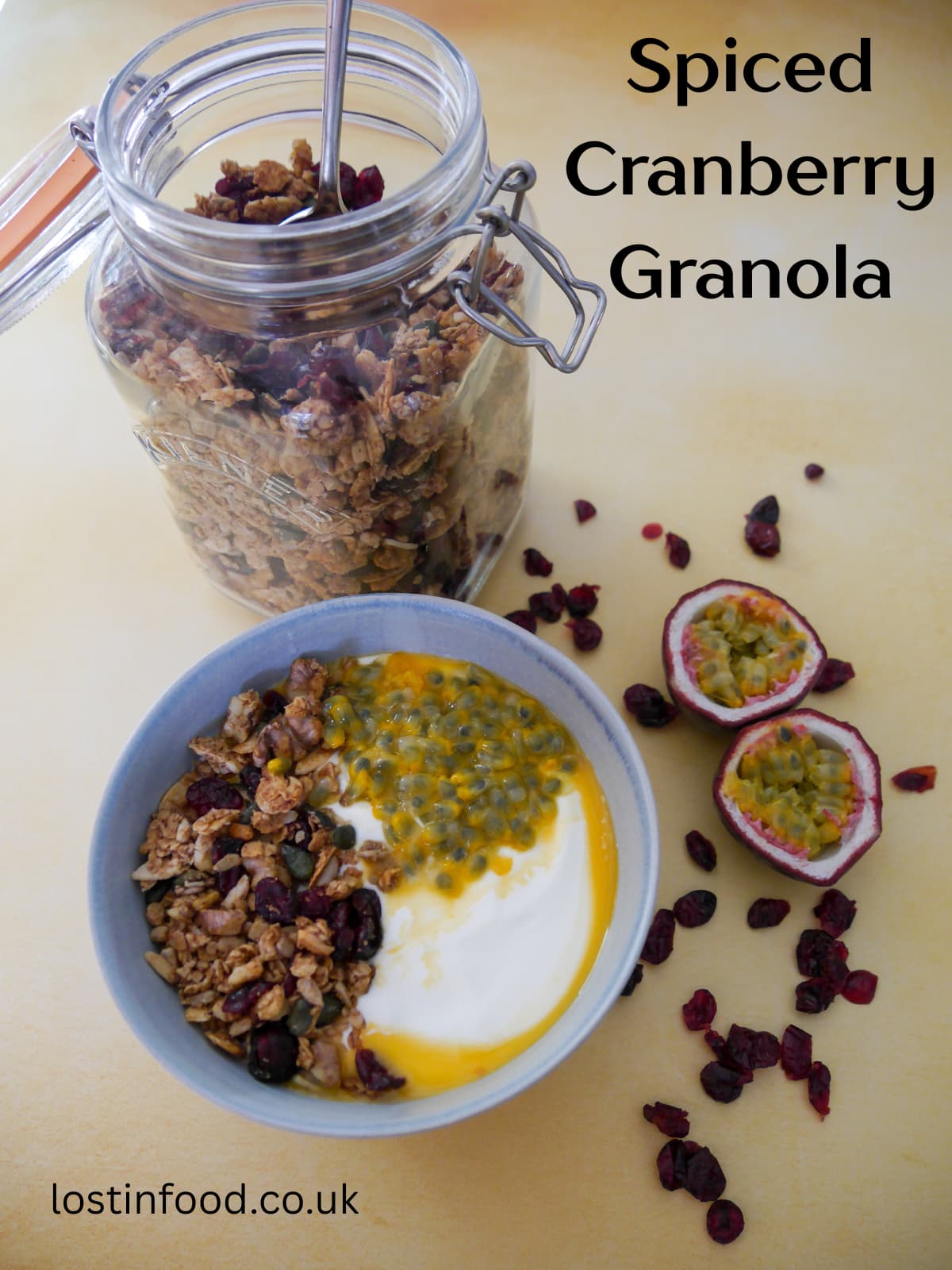 Pinnable image with recipe title and a bowl of Greek yogurt topped with spiced cranberry granola and passionfruit with a jar of granola set alongside.