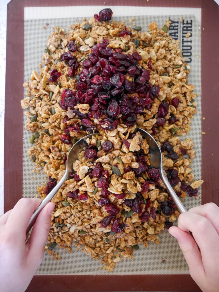 A lined baking sheet with cooked and cooled granola with dried cranberries being stirred through.
