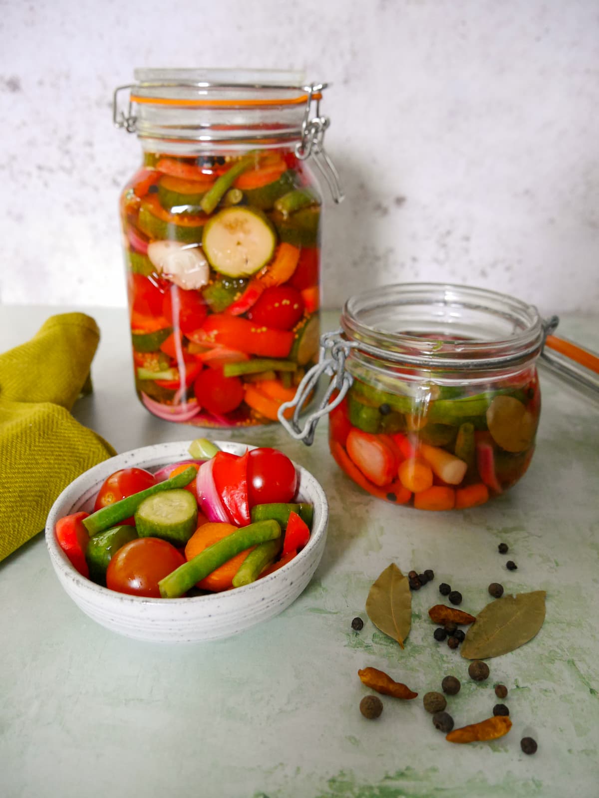 Jars of summer pickled vegetables with a bowl of vegetables and some dry spices set alongside.