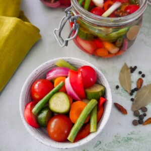 A bowl of summer pickled vegetables, with a large jar of pickled vegetables and some dry spices set alongside.