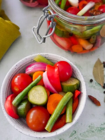 A bowl of summer pickled vegetables, with a large jar of pickled vegetables and some dry spices set alongside.
