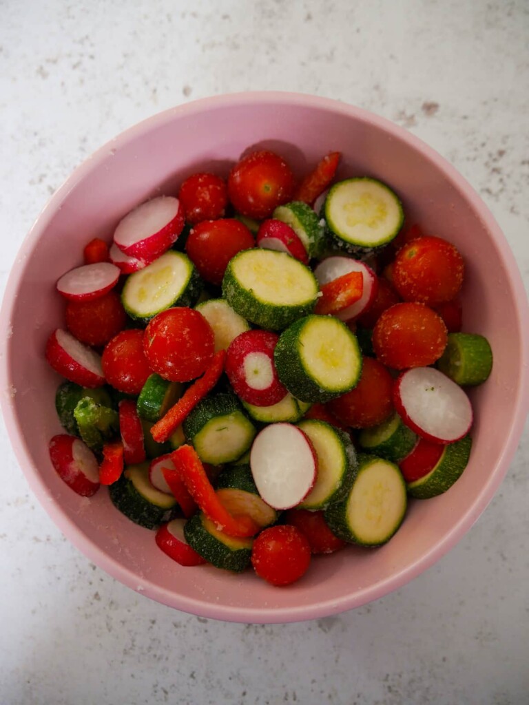 Sliced cucumber, courgette, radish, red pepper and whole cherry tomatoes in a bowl covered in sea salt.