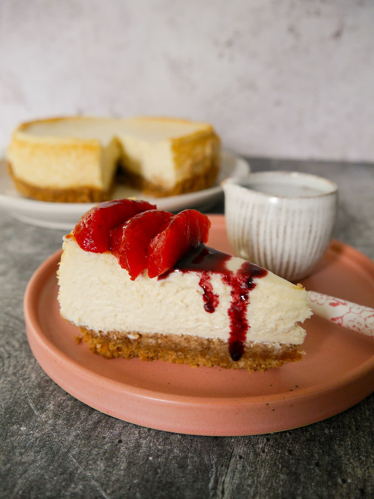 A plate with a slice of baked vanilla cheesecake, topped with some mulled plums and plum syrup, with the whole cheesecake set alongside.