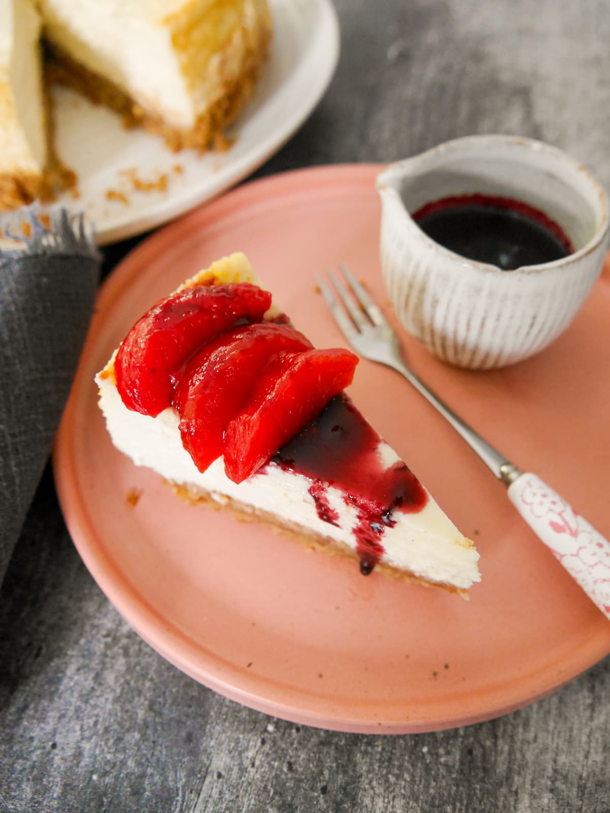 A plate with a slice of baked vanilla cheesecake, topped with some mulled plums and plum syrup, with the whole cheesecake set alongside.