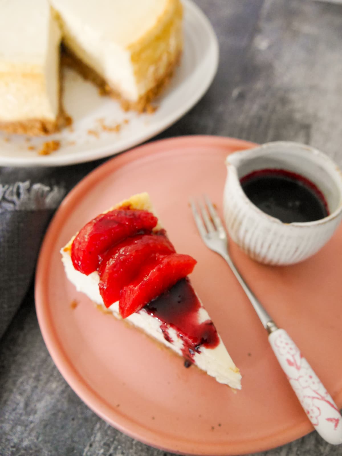 A plate with a slice of baked vanilla cheesecake, topped with some mulled plums and plum syrup, with the whole cheesecake set alongside.