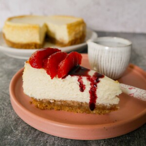 A plate with a slice of baked vanilla cheesecake, topped with some mulled plums and plum syrup, with the whole cheesecake set alongside.