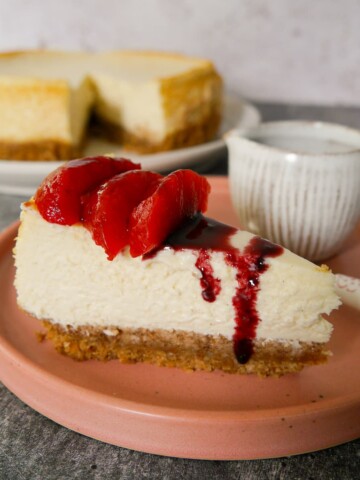 A plate with a slice of baked vanilla cheesecake, topped with some mulled plums and plum syrup, with the whole cheesecake set alongside.
