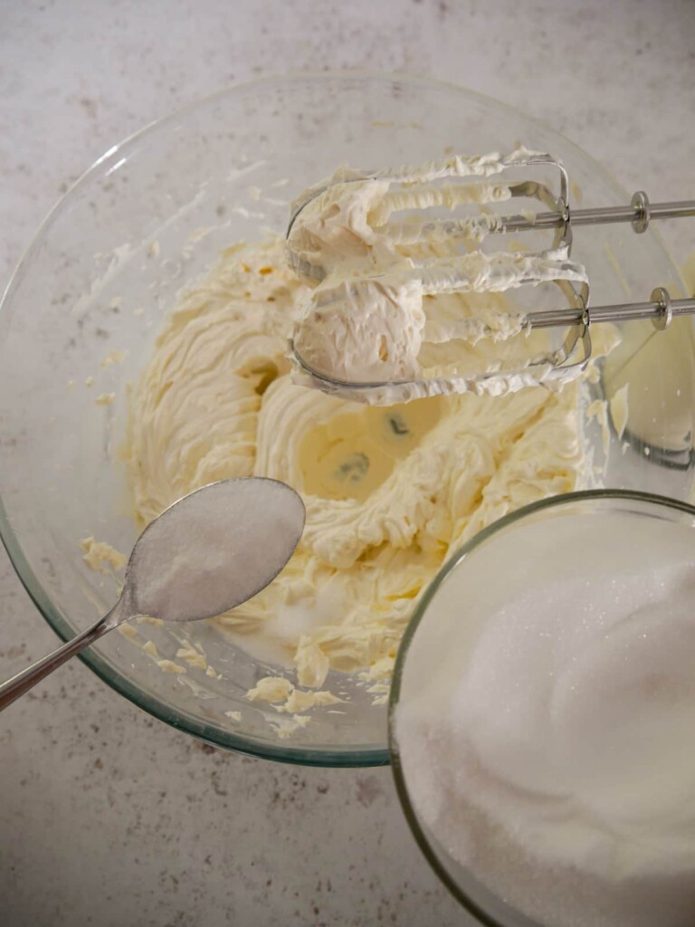A bowl of whipped cream cheese with spoonful's of caster sugar being added and whipped through.