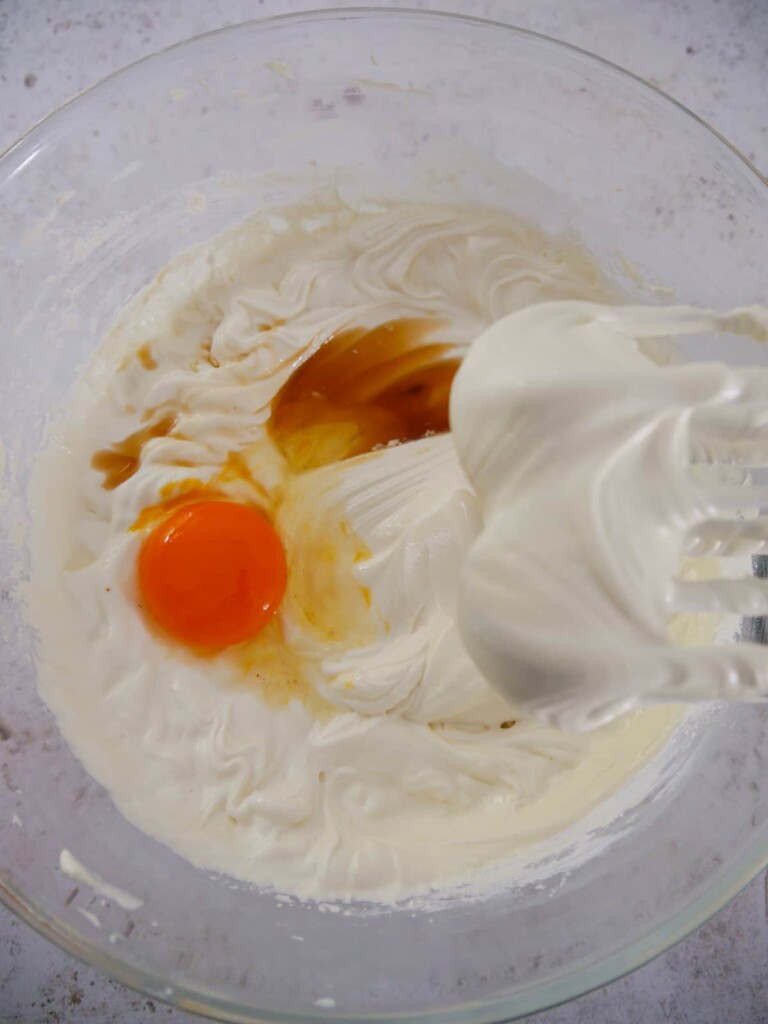 A bowl of whipped cream cheese and sugar with vanilla extract and eggs being whisked through.